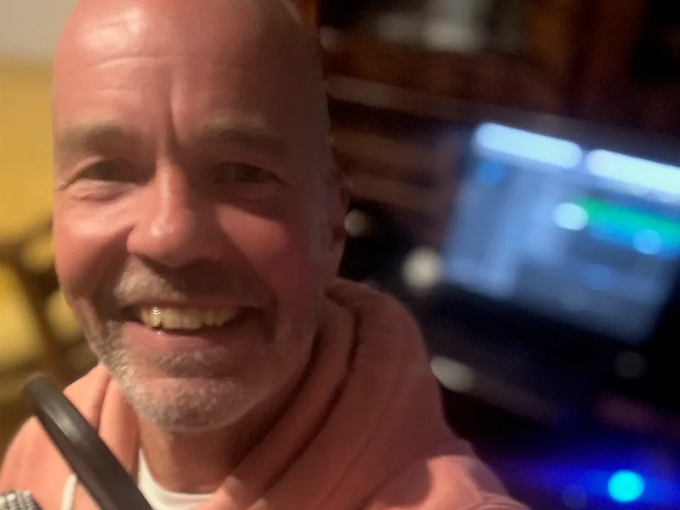 Henning lachend am Studio-Mikrofon, im Hintergrund unscharf ein Monitor mit DAW-Software.